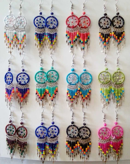 Twelve pairs of dreamcatcher-style dangle earrings displayed on a white wall. Each pair has silver-toned metal hooks, a circular woven center, and long beaded fringe in various colors including blue, red, turquoise, green, yellow, pink, and black.