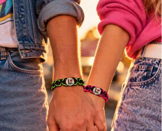 Two people holding hands, each wearing a braided friendship bracelet with a ceramic flat round charm engraved with I love 80s. One bracelet is black and green, and the other is pink and black.