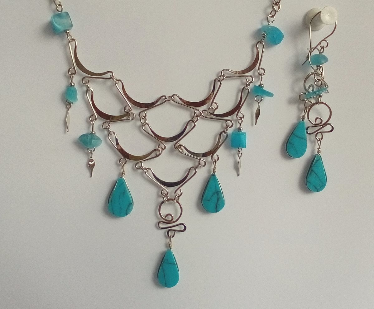 A jewelry set featuring a statement necklace and matching drop earrings with turquoise-colored teardrop beads and silver-toned metal chains and swirls, displayed against a plain light background.