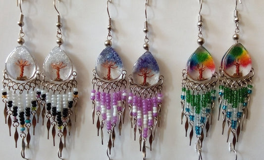 Three pairs of peruvian handmade dangling earrings. Each pair has a teardrop-shaped resin top with tree-of-life motifs, silver-tone metal frames, and beaded fringe in different color combinations: white/black, blue/purple, and rainbow/green.